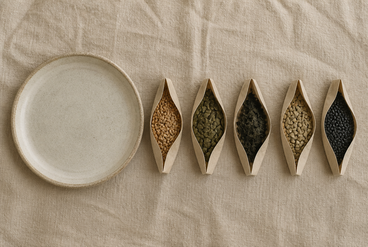 An empty stoneware plate beside five paper vessels holding wholegrains, seeds, seaweed flakes and lentils