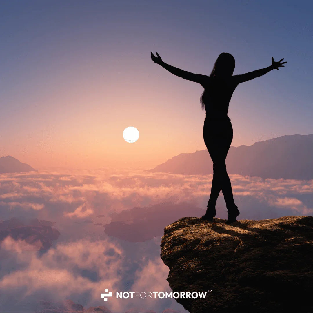 Silhouette of woman with arms raised on mountain cliff at sunrise above clouds