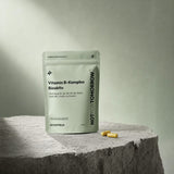 كيس NOTFORTOMORROW Vitamin B-Komplex Bioaktiv موضوع على منصة حجرية مع كبسولتين كريميتين أمامه