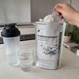 NOTFORTOMORROW Kreatinmonohydrat 500 g pose, en hånd løfter en skefuld pulver, ved siden af står en shaker og et glas vand