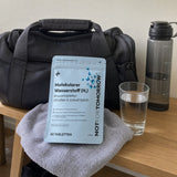 NOTFORTOMORROW Molecular Hydrogen tablet pouch on a towel by a gym bag, with a glass of water and a sports bottle