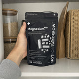 NOTFORTOMORROW Magnesium 7-in-1 zwarte zak met 120 capsules in één hand op een nette plank in de voorraadkast