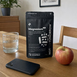 NOTFORTOMORROW Magnesium 7-in-1 zwarte zak met 90 capsules op een houten tafel met een glas water, een telefoon en een appel