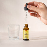 Hand houdt de glazen maatpipet met heldere olie boven het open amberkleurige NOTFORTOMORROW Vitamine D3 + K2 flesje